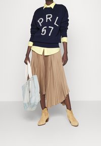 Navy sweatshirt with white "PRL 67" lettering, layered over a light yellow collared shirt. Pleated beige skirt, tan suede ankle boots, and light tote bag.