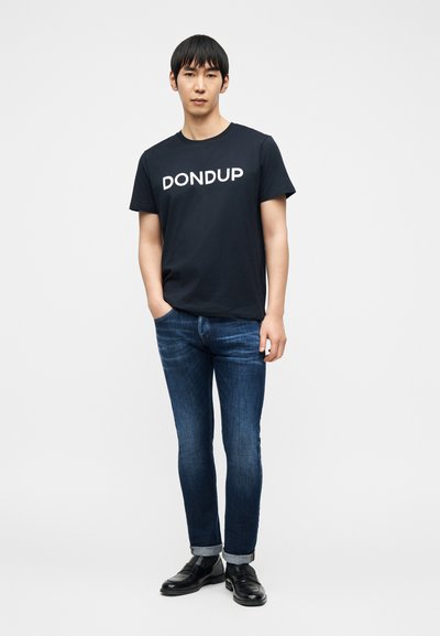 Νεαρός με μαύρα μαλλιά που φοράει μαύρο μπλουζάκι "DONDUP", μπλε τζιν με γυρισμένα μανίκια και μαύρα δερμάτινα παπούτσια, στέκεται με το ένα χέρι στην τσέπη.
