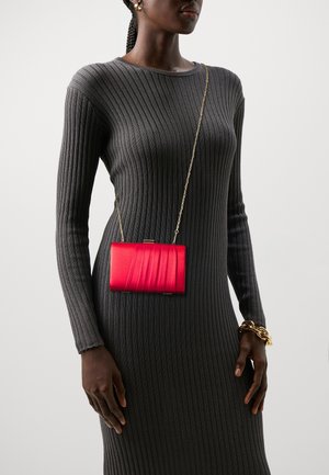 Pochette métallique rouge avec un design en vagues texturé, dotée d'une chaîne dorée. Portée sur une robe grise ajustée à côtes.