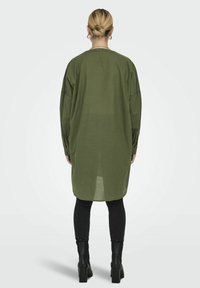 Chemise longue vert olive en tissu léger, avec une coupe ample, des épaules tombantes et des poignets boutonnés, associée à un legging noir et des bottines.