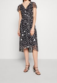 Černé midi šaty s květinovým vzorem, průhlednými krátkými rukávy, výstřihem do V a vázáním v pase. Obsahují bílé květinové vyšívání a splývavou sukni.