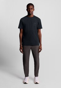 Zwarte T-shirt met korte mouwen en een vierkant logo op de borst, gecombineerd met donkergrijze tapered broek en lichte sneakers.