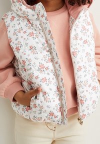 Biała bezrękawnik z nadrukiem kwiatowym z różowymi akcentami, z zamkiem błyskawicznym, miękką teksturą i stójką. Nosi się go na różową bluzkę.