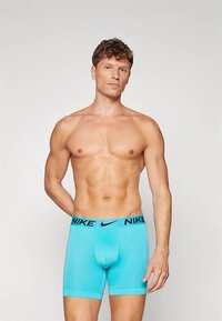 Izmos férfi egy sima háttér előtt, világoskék Nike boxer alsóneműt visel, a derékpánton a "NIKE" felirattal és a swoosh logóval.