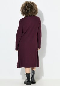 Langarmkleid aus tiefburgunderfarbener Strickware mit hohem Halsausschnitt und Seitenschlitzen. Kombiniert mit schwarzen knöchelhohen Stiefeln mit dicker Sohle.