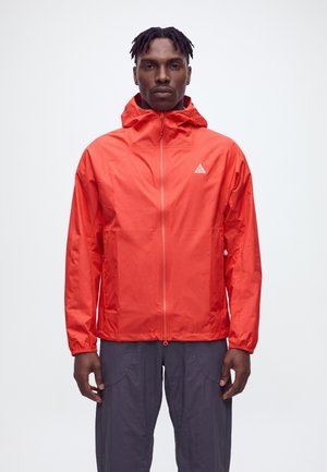 Un homme se tient face à l’objectif, vêtu d’une veste à capuche rouge vif zippée et d’un pantalon foncé, sur un fond clair et uni.