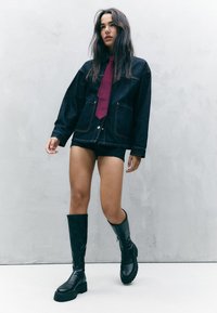 Džínová bunda s velkými kapsami, doplněná vínovou kravatou, černými kraťasy a černými vysokými botami s robustními podrážkami. Detaily texturované tkaniny.