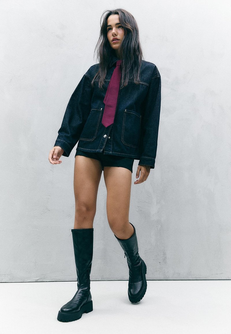 Džínová bunda s velkými kapsami, doplněná vínovou kravatou, černými kraťasy a černými vysokými botami s robustními podrážkami. Detaily texturované tkaniny.