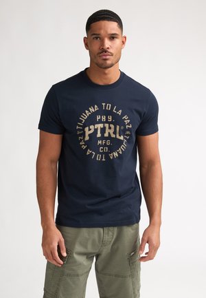 Mann trägt navyblaues T-Shirt mit rundem beigem Schriftzug und olivgrüne Cargohose, steht vor schlichtem Hintergrund.