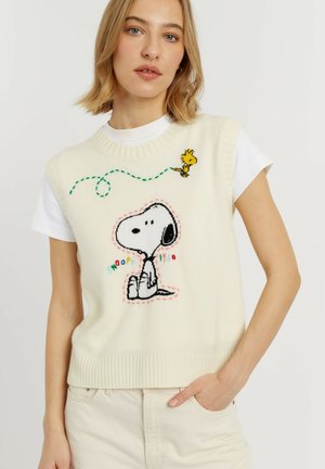 Vrouw draagt een wit gebreid vest met Snoopy en Woodstock cartoon en een wit shirt met korte mouwen, staat met hand in de zak.