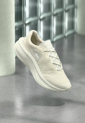 Λευκό αθλητικό παπούτσι Lacoste με διάτρητες πλευρές και παχιά σόλα που αιωρείται σε εσωτερικό χώρο πάνω σε γυαλισμένο τσιμεντένιο δάπεδο κοντά σε γυάλινους κυβόλιθους.