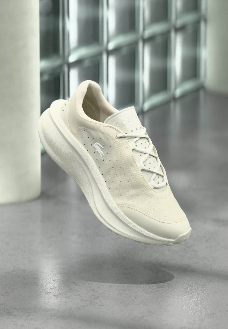 Λευκό αθλητικό παπούτσι Lacoste με διάτρητες πλευρές και παχιά σόλα που αιωρείται σε εσωτερικό χώρο πάνω σε γυαλισμένο τσιμεντένιο δάπεδο κοντά σε γυάλινους κυβόλιθους.