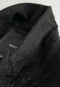 Černá tkanina s vzorem a jemnou texturou a leskem, se skládacím límcem a štítkem se značkou "NEXT."
