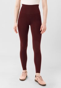 Leggings burgundi a vita alta realizzati in tessuto elasticizzato, design aderente con texture liscia, abbinati a scarpe da ginnastica rosa chiaro.