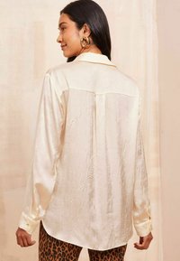 Camicia di satin color crema con finitura tessurizzata, maniche lunghe e orlo arrotondato. Presenta un colletto classico e polsini con bottone.
