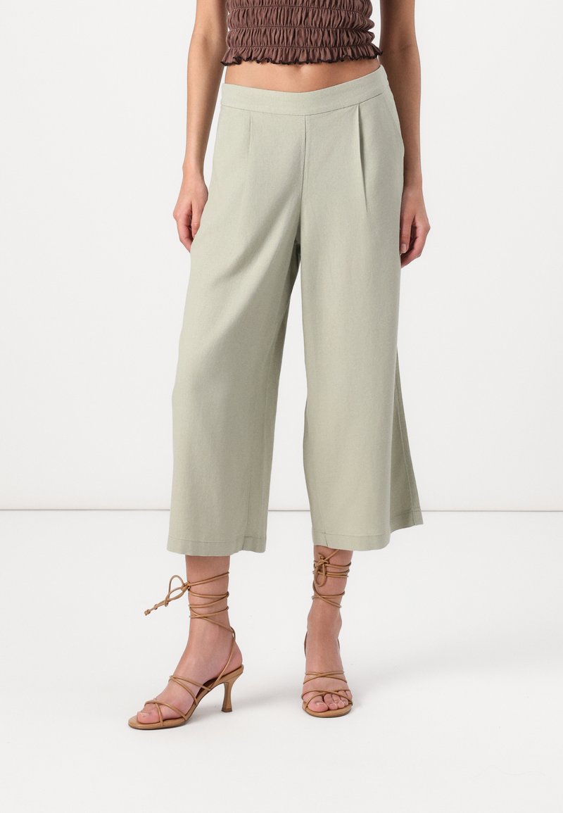 Pantalons larges vert clair avec des plis sur le devant, texture douce, taille haute. Talons bruns à lanières avec un talon fin, orteils découverts.