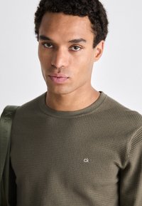 Maglione verde a trama con scollatura rotonda, caratterizzato da un piccolo logo "ck" bianco sul lato sinistro del petto e una tracolla coordinata.