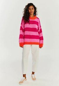 Pletený oversize svetr s výraznými růžovými a fuchsiovými pruhy, zvýrazněný oranžovým žebrovaným límcem. V kombinaci s bílými kalhotami a světlými teniskami.