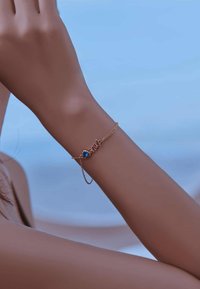 Pulsera de cadena de oro con un charm en forma de corazón azul y la palabra "amor" en una fuente cursiva, mostrada en un brazo sobre un fondo azul.