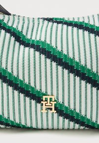 Zielona i biała pleciona torba w paski z złotym emblematem i ciemnoniebieskimi akcentami. Posiada teksturowaną powierzchnię i prostokątny kształt.