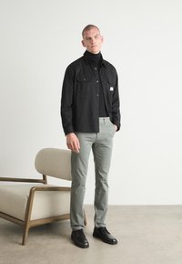Cămașă neagră cu nasturi, dotată cu două buzunare pe piept, asociată cu pantaloni slim-fit de culoare verde deschis și pantofi negri, pe un fundal neutru.