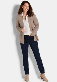 Frau lächelt, trägt eine beigefarbene Strickjacke, eine weiße Bluse, eine dunkelblaue Hose und beigefarbene Stiefeletten und steht vor einem schlichten hellen Hintergrund.