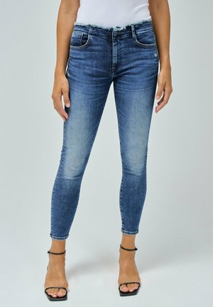 Femme portant un jean bleu skinny taille haute avec une ceinture effilochée et des talons noirs à fines brides, debout devant un fond clair uni.