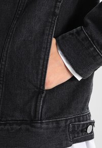 Czarna kurtka dżinsowa z przednią kieszenią, kontrastowymi szwami i detalem z guzikiem przy mankiecie. Tkanina ma teksturowane, matowe wykończenie.
