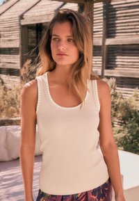 Beige tanktop med scoop-neck og ribbet tekstur samt dekorative hvide syninger langs halsudskæringen. Kombineret med farverige mønstrede shorts.