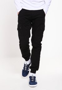 Man i svarta cargobyxor med knäppta fickor och marinblå Adidas-sneakers som går framför en enfärgad vit bakgrund.