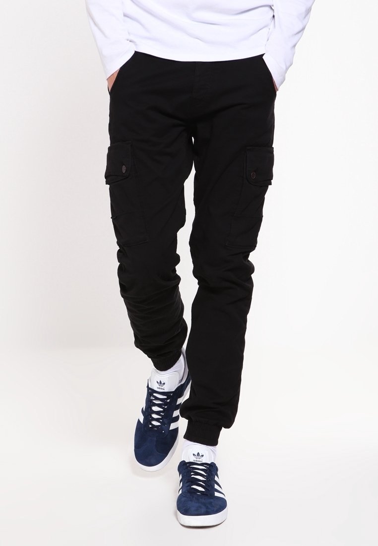 Man i svarta cargobyxor med knäppta fickor och marinblå Adidas-sneakers som går framför en enfärgad vit bakgrund.