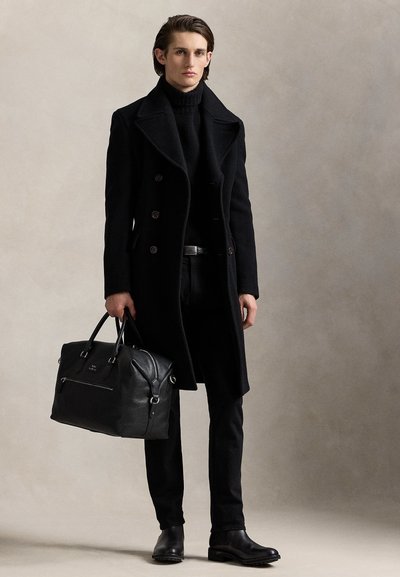 Jeune homme portant un manteau noir croisé, un col roulé noir, un pantalon noir et des bottes, tenant un grand sac de voyage en cuir noir.