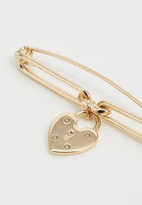 Spilla di sicurezza in oro con un ciondolo a forma di cuore caratterizzato da un design a lucchetto e cinque rivetti decorativi. Texture liscia e lucida su tutta la superficie.