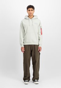 Ανοιχτό πράσινη έμπνευση με μπροστινή τσέπη, κόμπο και μανίκι από ύφασμα· σε συνδυασμό με φαρδιά ελαιόλαδα παντελόνια και μαύρα-λευκά αθλητικά παπούτσια.