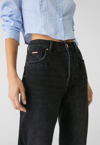 Jeans de denim negro de talle alto con corte relajado, que presentan herrajes en tono cobre, bolsillos frontales y una textura sutil.