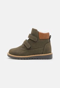 Bottines en daim vert avec deux bandes Velcro, accent de talon brun contrastant, doublure en fourrure blanche et semelle en caoutchouc noir robuste.