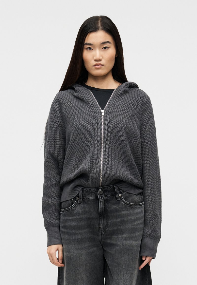 Jeune personne aux longs cheveux noirs portant un sweat à capuche en maille gris foncé zippé sur un t-shirt noir et un jean noir délavé, debout devant un fond blanc.