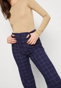 Kvinna iklädd en beige långärmad topp och marinblå rutiga byxor med krage och bältesöglor, med händerna vilande vid midjan.