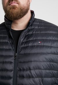Svart puffjacka med horisontell quiltstruktur, hög krage och dragkedjestängning. Har en liten röd broderad logotyp på bröstet.