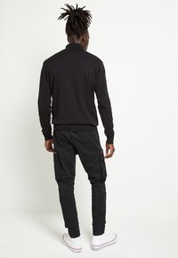 Black knitted sweater with a high collar, worn with black cargo pants featuring multiple pockets, and white sneakers with red accents.