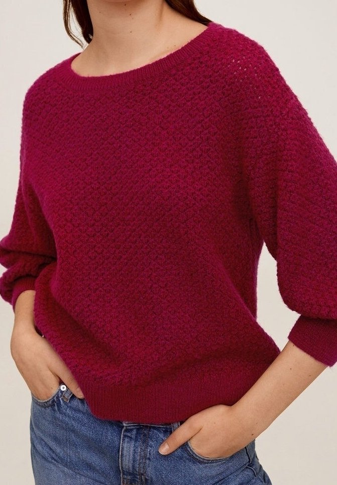 Różowy sweter dziergany z teksturowanym wzorem, z okrągłym dekoltem i prążkowanymi mankietami. Stylizowany z jasnoniebieskimi jeansami o luźnym kroju.