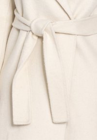 Primo piano di una cintura annodata su un morbido cappotto di lana color crema con uno stile di nodo avvolto.