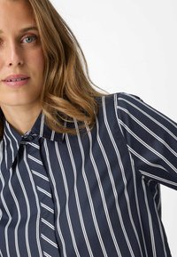 Camicia a bottoni a righe blu navy con strisce verticali bianche, colletto e tessuto morbido. Decorata con righe di larghezza variabile.