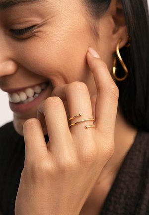 Gold rings with a minimalist design featuring smooth, curved shapes. A hand rests gently, showcasing two thin bands with small spherical accents.