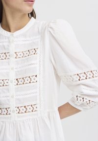 Camicia bianca con chiusura a bottoni, presenta inserti in pizzo e maniche a sbuffo. Realizzata in tessuto leggero con una finitura testurizzata.