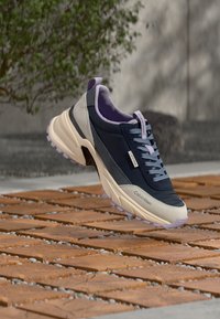 Ναυτικό μπλε και μπεζ sneaker Calvin Klein με ανοιχτό μοβ λεπτομέρειες, αιωρείται πάνω από ξύλινο δάπεδο με πλακάκια σε  εξωτερικό χώρο κοντά σε πράσινο φυτό.