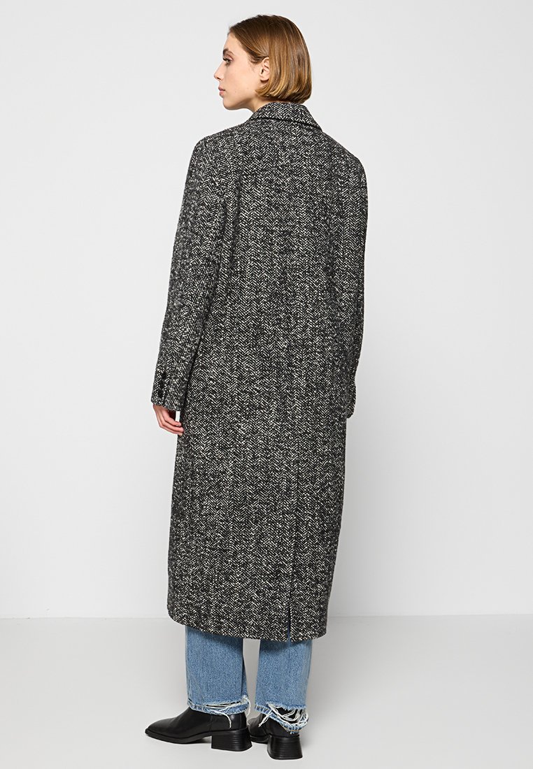 Lang, grijs houndstooth overjas met een gesneden kraag en achteropening, gecombineerd met rafelige lichtblauwe jeans en zwarte enkellaarsjes.