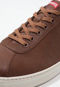 Sneaker de cuero marrón con puntera redonda, costuras visibles y cordones marrones. Presenta una suela color crema y una etiqueta de marca roja en la lengua.