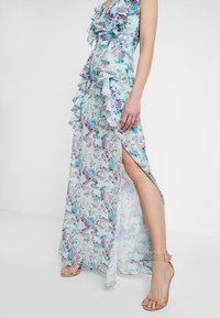 Femme portant une robe maxi florale bleu clair avec des volants et une fente haute, associée à des sandales à talons aiguilles nud à brides autour de la cheville.