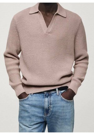 Man draagt een lichtbruine trui met een gebreide kraag, lange mouwen, en een blauwe spijkerbroek, handen in de zakken, tegen een effen achtergrond.